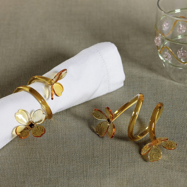 The Botanica Napkin Ring set of 4 by Zodax features gold and enamel butterfly designs displayed on a beige tablecloth, with one ring elegantly holding a white napkin, while a glass adorned with pink flowers in the background enhances this sophisticated botanical table decor.