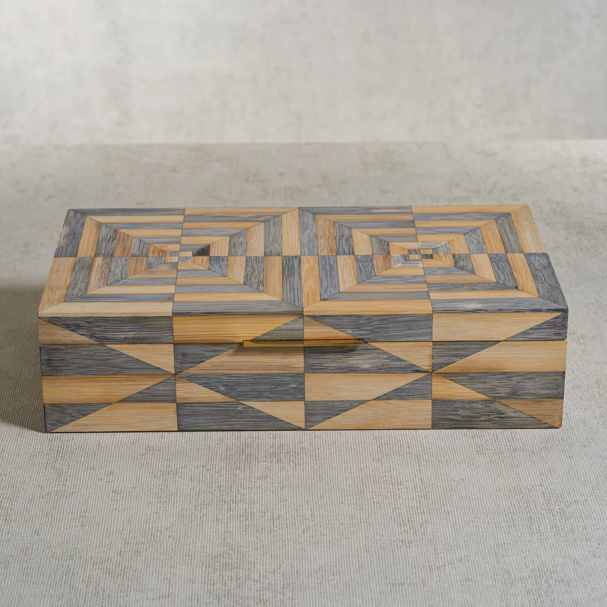 The Zodax Monarch Bamboo Box w/ Brass Trim features a rectangular silhouette adorned with intricate geometric patterns in gray and tan hues, forming concentric diamonds and triangles across the lid and sides, and is elegantly finished with subtle brass trim. Shown here resting on a light textured surface.