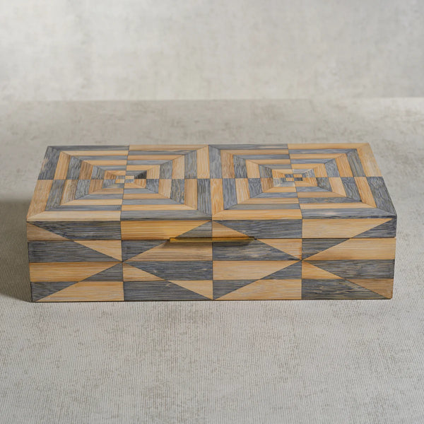 The Zodax Monarch Bamboo Box w/ Brass Trim features a rectangular silhouette adorned with intricate geometric patterns in gray and tan hues, forming concentric diamonds and triangles across the lid and sides, and is elegantly finished with subtle brass trim. Shown here resting on a light textured surface.