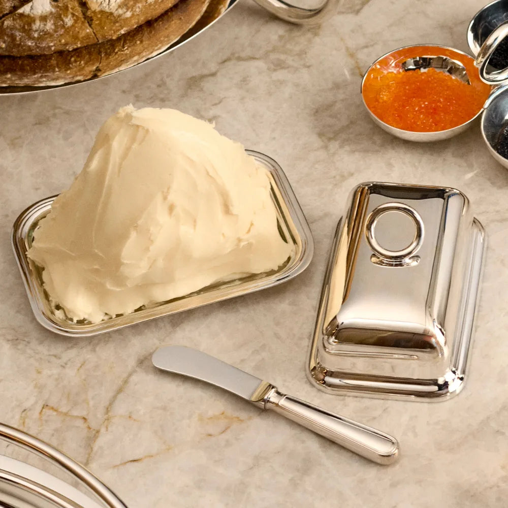 A Christofle Vertigo Silver-Plated Butter Dish with Lid featuring a mound of butter on the tray and a butter knife resting on a marble surface.