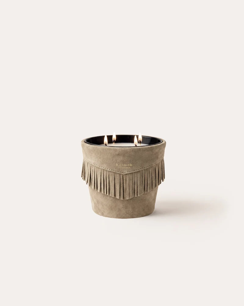 A Baobab Calamity Jane Candle - The Plains, featuring three wicks in a mineral taupe leather, light brown fringed container with a single flame lit.