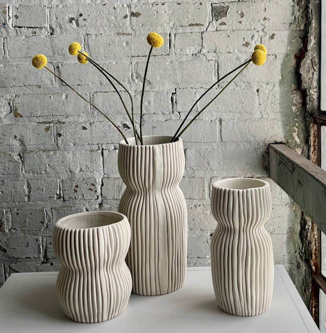 Three handmade in USA, cream-colored, ribbed Round Curvy Vase Cream vases of varying heights, with thin stems topped with yellow billy button flowers in the center vase.