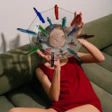 A person in a red outfit sits on a green couch, holding the Yakush Japanese Winter Bowl - Neon, a clear hand-blown glass bowl with colorful clothespins attached to its edge, obscuring their face.