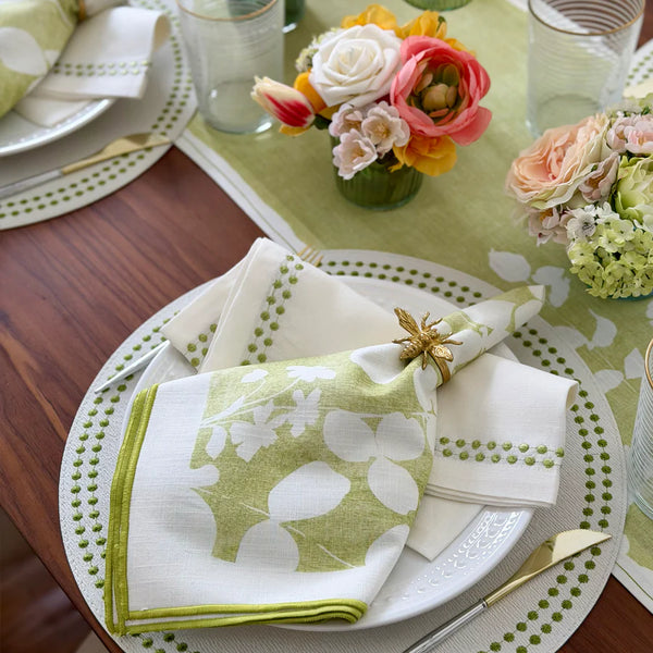 A set table with white plates, Bodrum Fleur de Lin Napkins in green, a gold bee napkin ring, and a bouquet of pink and white flowers on a green table runner.