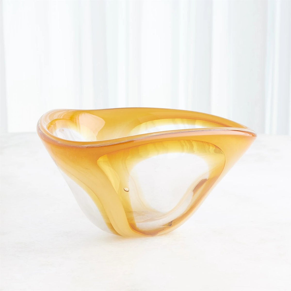 The Enigma Bowl - Amber by Global Views / Surya, a sculptural art glass piece featuring abstract, flowing curves and a warm amber hue, rests on a light surface against a softly lit, white background.