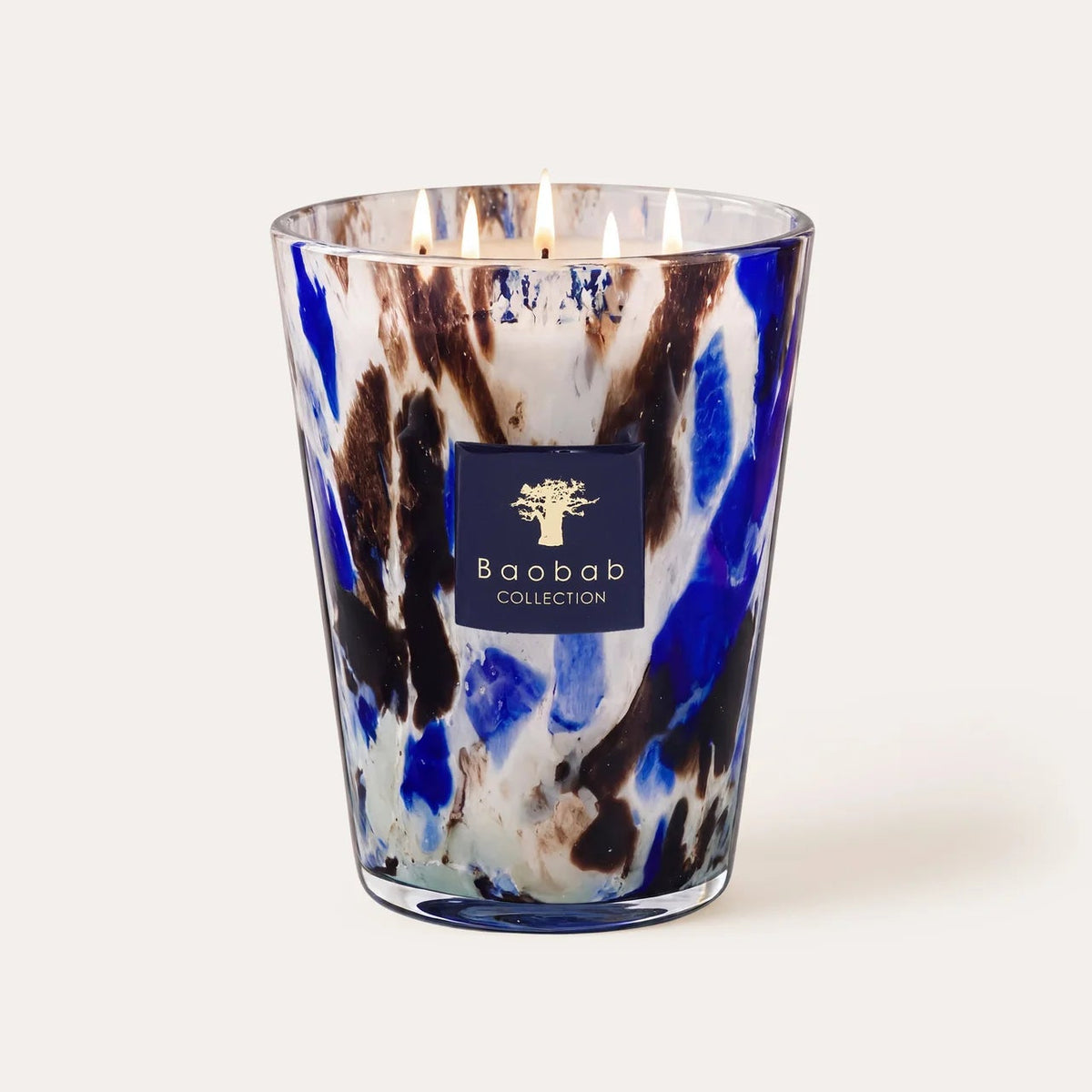 A lit Baobab Halona Candle Blue World, presented in a hand-blown glass holder with blue, white, and brown marbled patterns and the Baobab logo on the front. This woody fragrance candle blends notes of rosemary, cedarwood, and vetiver to create a sophisticated ambiance.