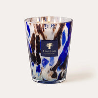 A lit Baobab Halona Candle Blue World, presented in a hand-blown glass holder with blue, white, and brown marbled patterns and the Baobab logo on the front. This woody fragrance candle blends notes of rosemary, cedarwood, and vetiver to create a sophisticated ambiance.