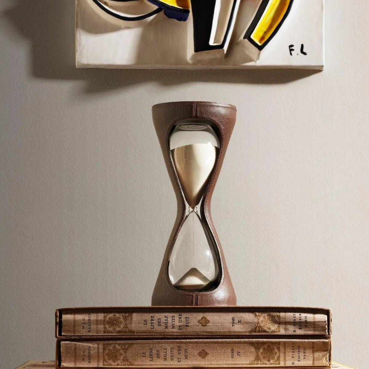 The Mirage Luxury Leather Hourglass by Assouline, featuring white sand and a leather exterior, is displayed atop two vintage books beneath vibrant abstract wall art, standing out against the light-colored wall.