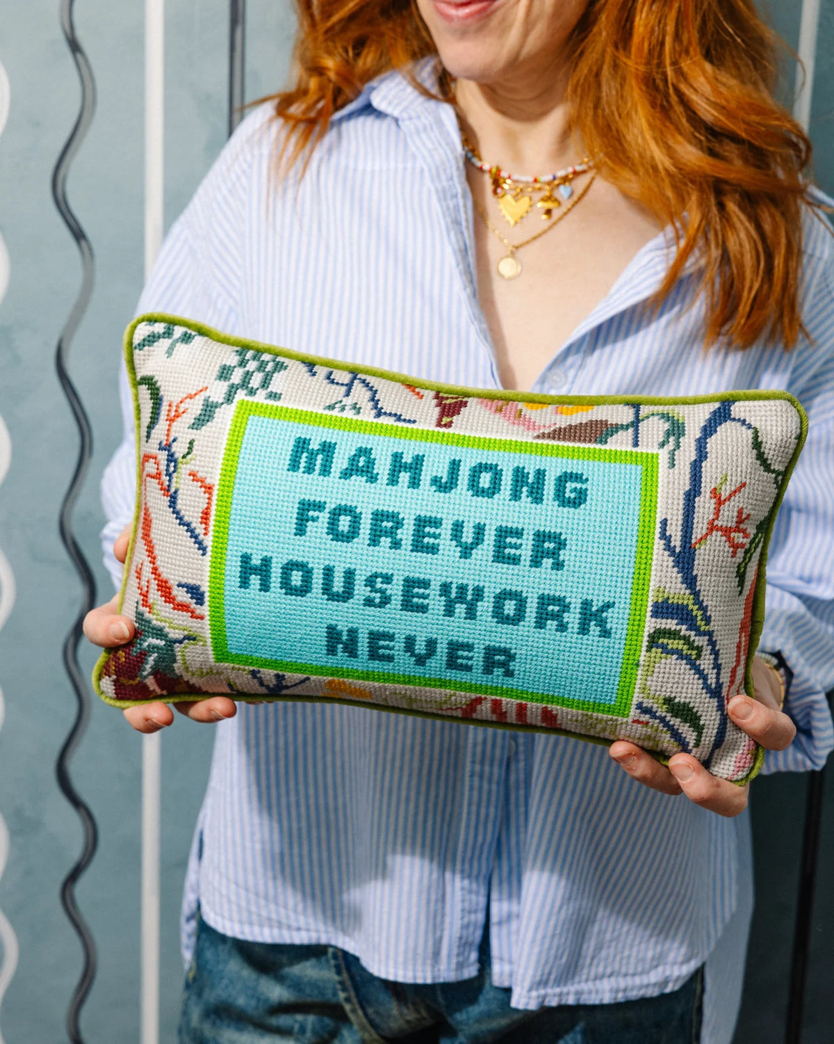 A woman with red hair holds the Furbish Needlepoint Pillow - Mahjong, which reads "MAHJONG FOREVER HOUSEWORK NEVER" in blue text—an ideal gift for Mahjong fans who love this classic game.