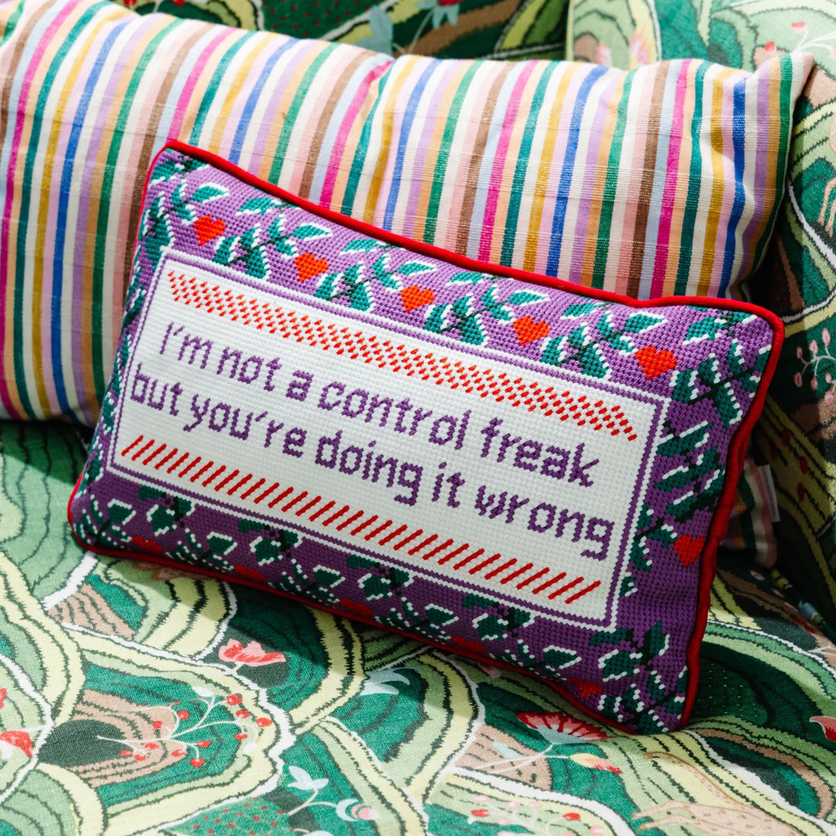 The Furbish Needlepoint Pillow - Control Freak features colorful embroidery that reads, "I'm not a control freak but you're doing it wrong," with floral patterns, shown on patterned fabric next to a striped pillow.