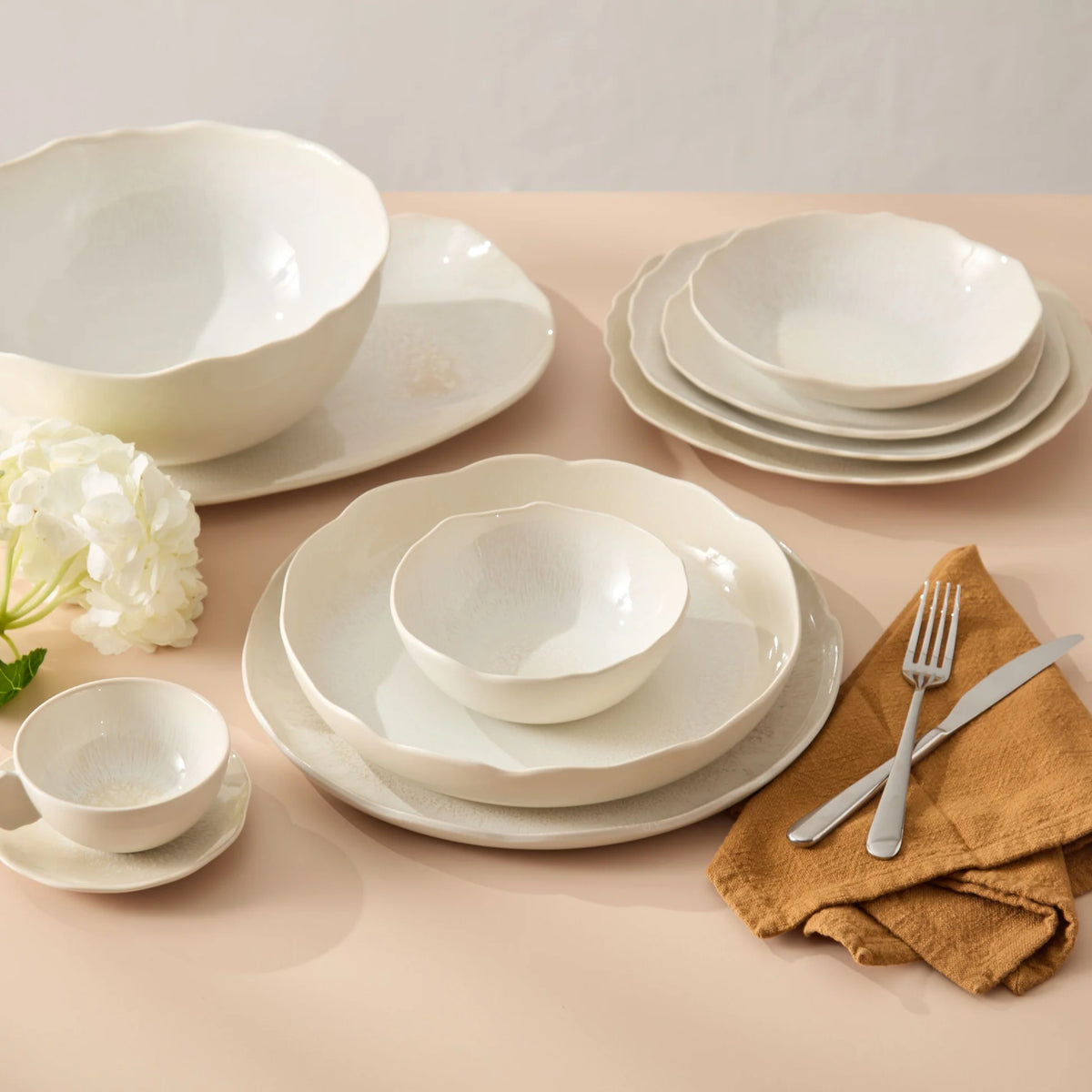 Cream-toned ceramic pieces from the Jars - Rosenthal Plume White Dinnerware Pearl collection are artfully arranged, featuring plates, bowls, and a teacup with matching saucer on a beige surface. Each piece is enhanced by delicate crystallised enamels that introduce subtle texture. A white flower and gold napkin with silverware complete this refined setting.