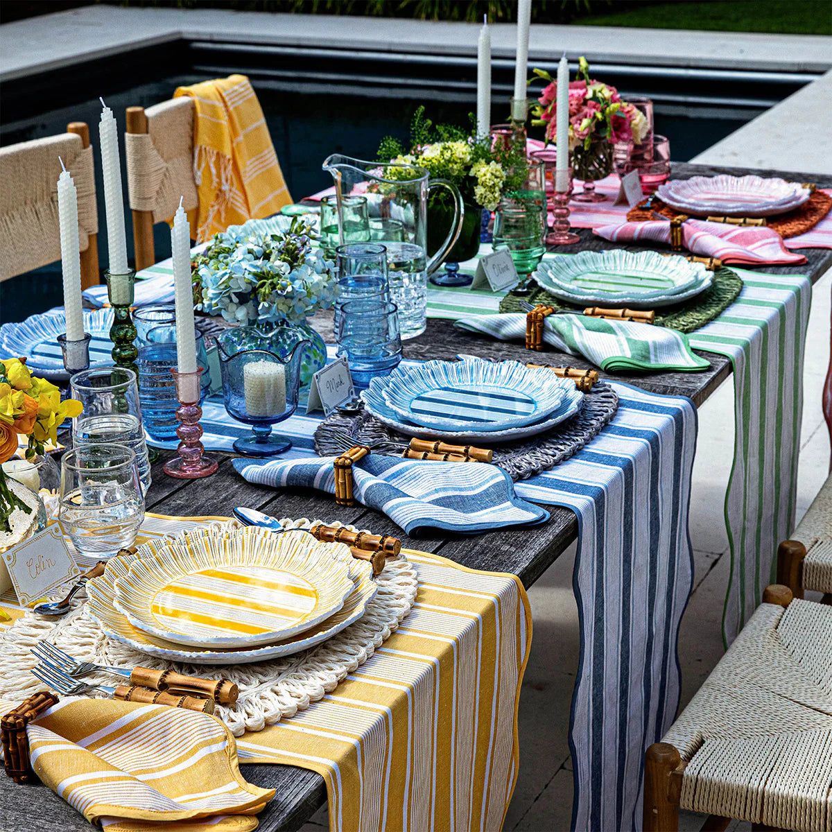 A festive poolside table is set with Juliska's Cabana Stripe Melamine Dinnerware Set of 4 in Coral, featuring striped plates, napkins, and table runners, along with candles, glassware, flowers, and place cards for a vibrant outdoor gathering.