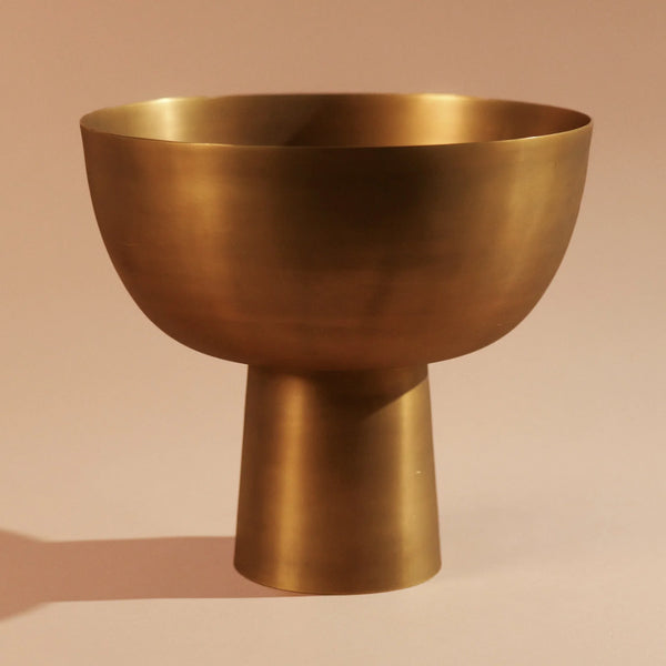 The Kara Brass Footed Bowl by Dumae stands elegantly against a warm beige background, its handcrafted metal form featuring a wide, rounded top and a narrow, cylindrical base—making it a stunning piece of artisan home decor.