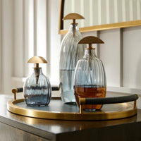 Three Karis Decanter pieces by Arteriors, each with ribbed optic glass and gold dome stoppers, rest on a round antique brass tray atop wood, backed by a mirror and white paneled wall.
