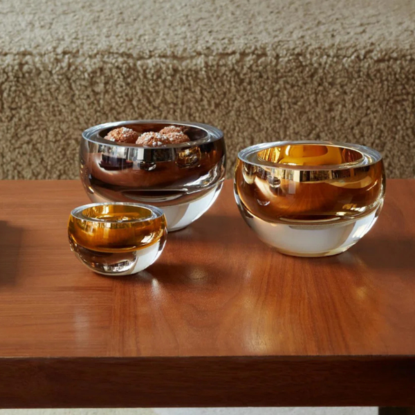 Three decorative glass bowls with an amber tint sit on a wooden table. The largest, the LSA Hostess Bowl- Platinum Large, is expertly crafted from mouth-blown glass in Poland and holds truffles, while the other two remain empty. A textured beige surface appears in the background.