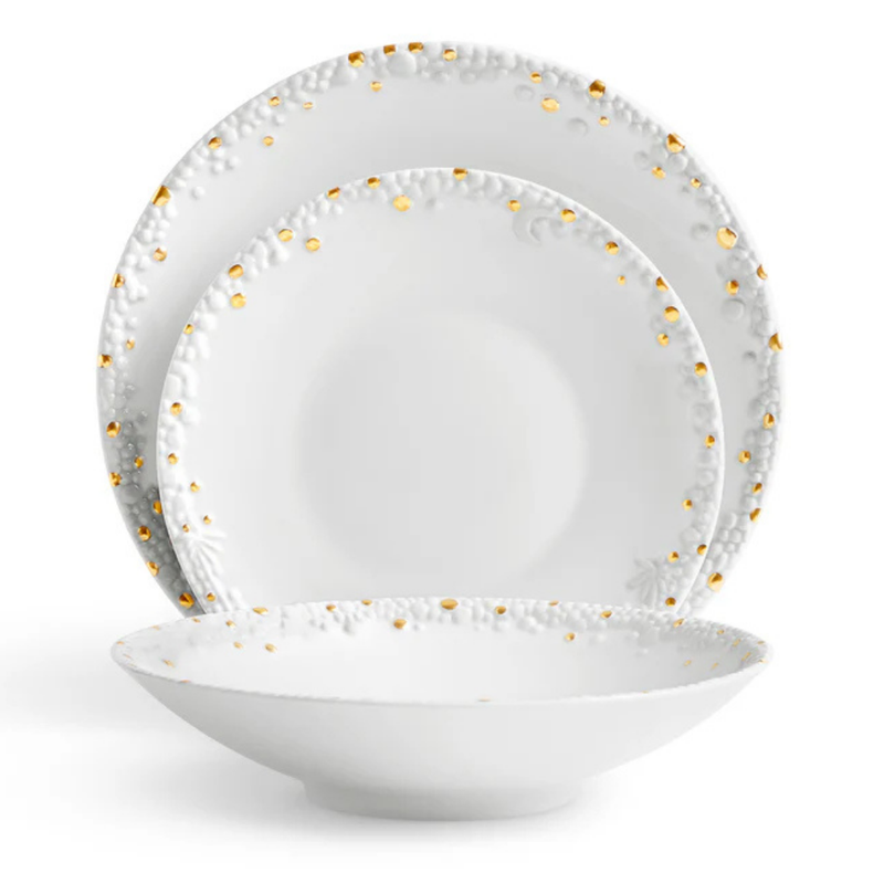 Three white Haas Mojave Dinnerware Gold pieces by L'Objet—each with embossed patterns and gold dot accents—are arranged: a dinner plate at the back, salad plate in the middle, and a shallow bowl at the front.