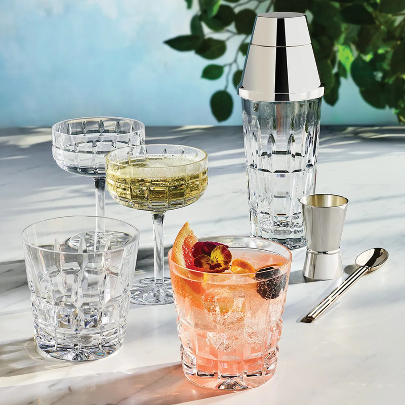The Saint-Louis Manhattan Barware set features a coupe and two tumblers—one with a yellow cocktail, one empty, and one with a pink drink and garnishes—plus a metal shaker, jigger, and spoon on marble with leafy shadows.