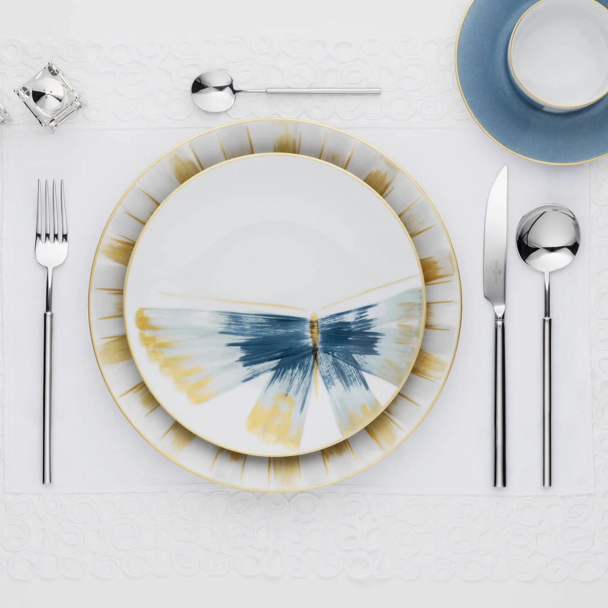 Place setting featuring Marie Daâge Belle De Nuit 1 porcelain plates with a blue and gold butterfly-like pattern, a blue teacup, polished silverware, and a crystal napkin holder on a white lace placemat.