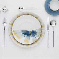 Place setting featuring Marie Daâge Belle De Nuit 1 porcelain plates with a blue and gold butterfly-like pattern, a blue teacup, polished silverware, and a crystal napkin holder on a white lace placemat.