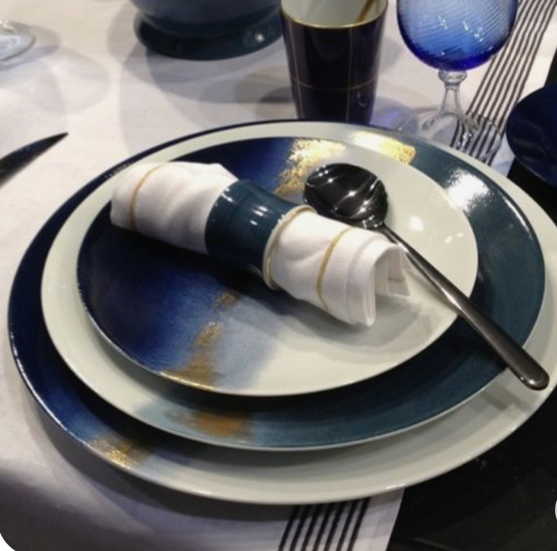 A table setting with Marie Daâge Horizon Gold Dinnerware, featuring blue and white gradient plates, a white napkin in a blue ring, and a spoon on top. Glassware and other French tableware appear in the background on a white tablecloth.