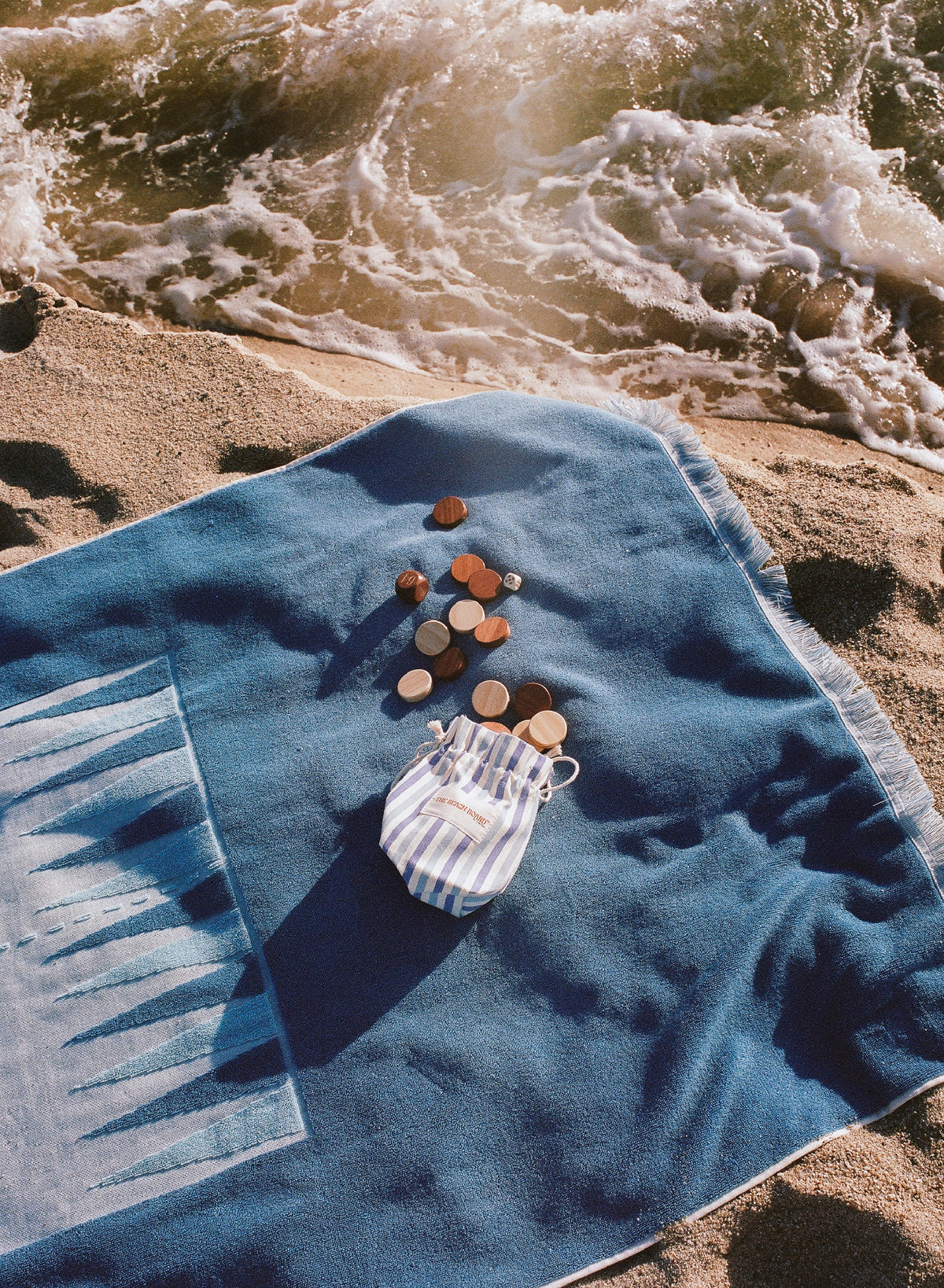 The G. McGee Southampton Beach Backgammon set rests on the sand near the shore, its striped drawstring pouch and scattered seashells atop, while sunlight glimmers off ocean waves in the background.