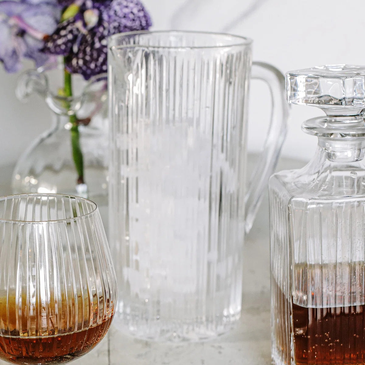 The Vietri Natalia Pitcher filled with ice, accompanied by a matching glass containing a brown liquid and a faceted decanter holding amber liquid, are arranged on a table. In the background, flowers in a vase appear softly blurred.