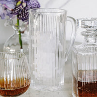 The Vietri Natalia Pitcher filled with ice, accompanied by a matching glass containing a brown liquid and a faceted decanter holding amber liquid, are arranged on a table. In the background, flowers in a vase appear softly blurred.