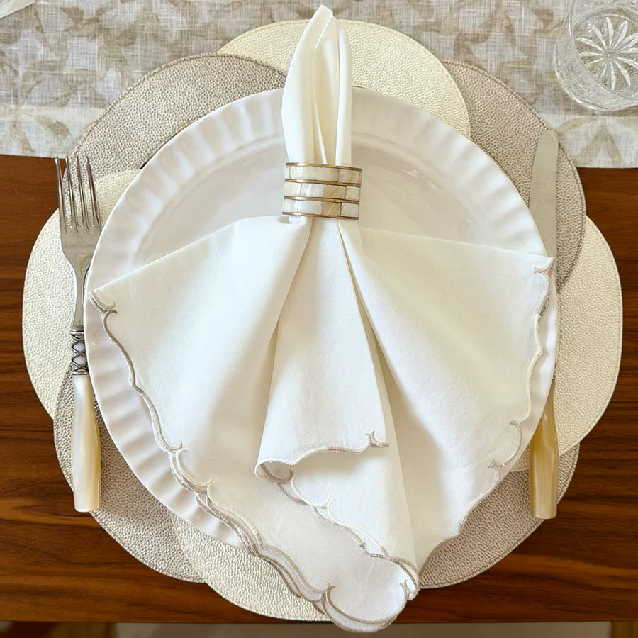 A white plate with a scalloped napkin from Bodrum's Firenze Napkin Set of 4 - Neutral, folded upright in a silver ring and placed between a fork and knife, rests on layered placemats atop a wooden table.