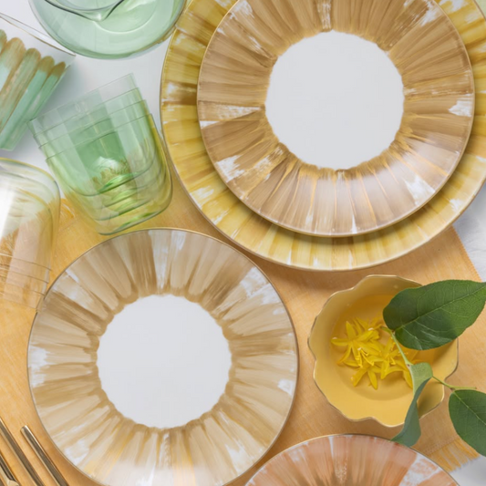 A flat lay of a table setting with Marie Daâge Panache luxury dinnerware, showcasing gold and white patterned plates, green glassware, and a small yellow dish adorned with yellow flowers and a green leaf.