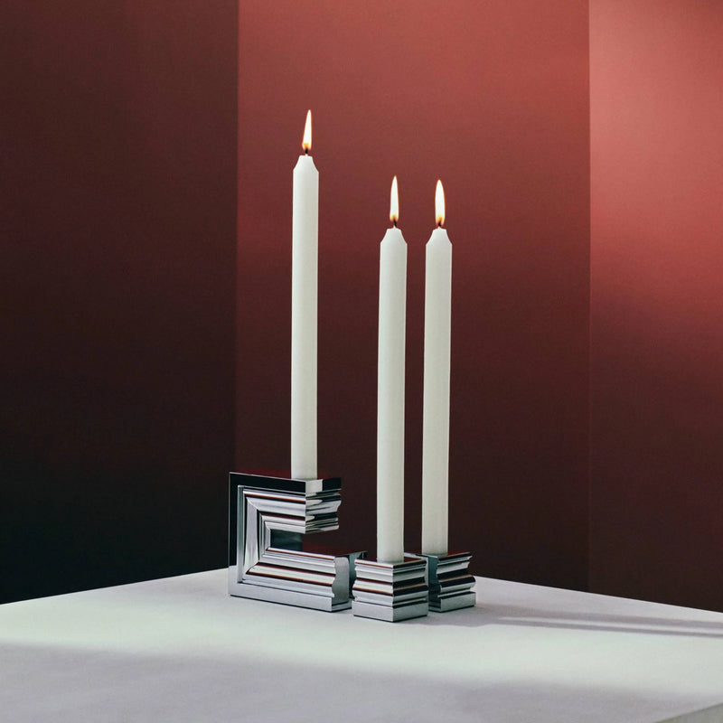 Three tall white candles are lit and placed in the Perspectives Chrome Candle Holder Large by Christofle, set on a white surface with a red gradient background.