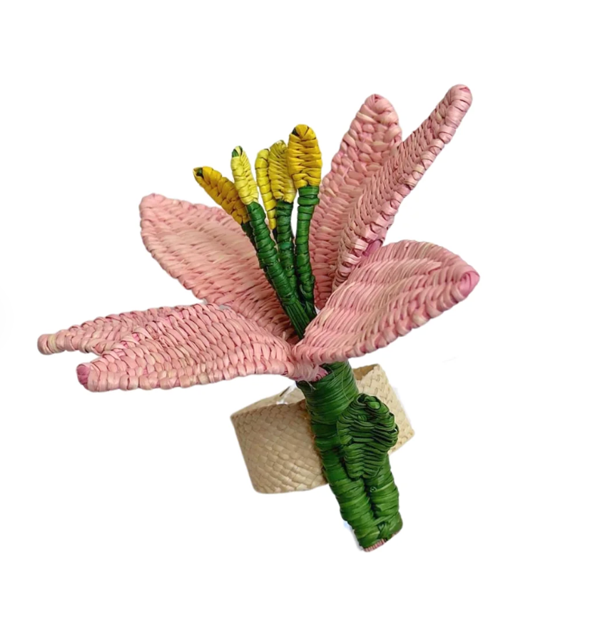 The Coro Cora Lily Napkin Ring Set of 4 in pink features flower-shaped, handwoven natural straw fibers with green and yellow accents, adding a charming touch to outdoor dining. Displayed on a white background.