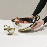 A person arranges the Anna New York Ring Fruit Bowl, a shiny stainless steel piece with gold accents filled with dragon fruit, as a centerpiece beside a matching smaller bowl holding strawberries on a white surface.