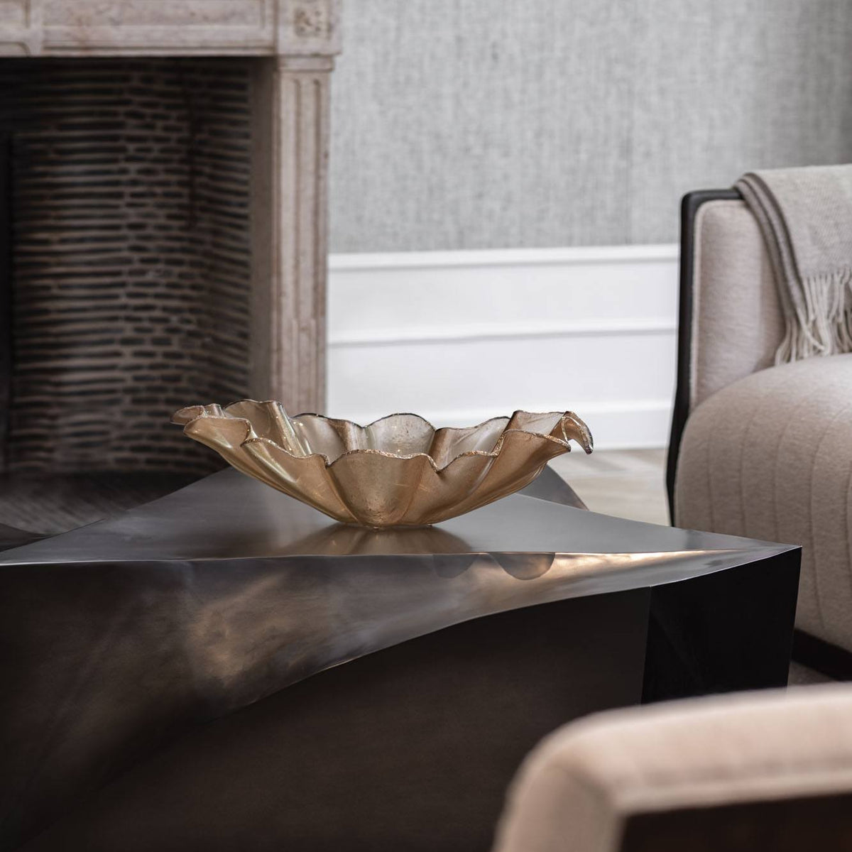 An Anenoma Bowl with copper and bronze trim by Gardeco sits on a dark, angular coffee table in a modern living room with a fireplace, a beige armchair, and a light gray wall.