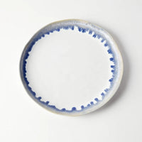 A round ceramic plate from Simon Pearce’s Burlington Dinnerware Pool collection showcases a white center accented by a blue, watercolor-inspired ring along its slightly irregular glazed edge, lending it an organic appearance when displayed on a white background.