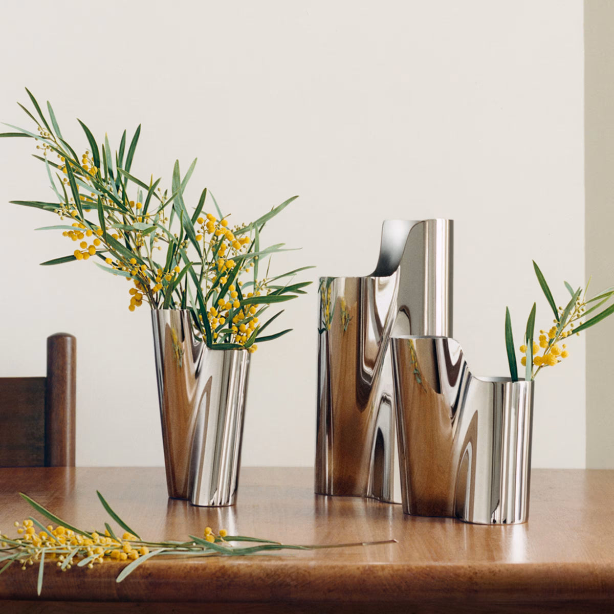 Three silver, wavy sculptural Lituus Vases by Georg Jensen, each filled with yellow flowers and green leaves, sit on a wooden table next to a wooden chair. Another sprig of flowers rests on the table. The background features a plain off-white wall.
