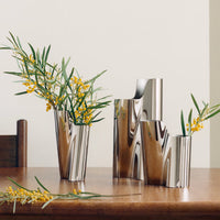 Three silver, wavy sculptural Lituus Vases by Georg Jensen, each filled with yellow flowers and green leaves, sit on a wooden table next to a wooden chair. Another sprig of flowers rests on the table. The background features a plain off-white wall.