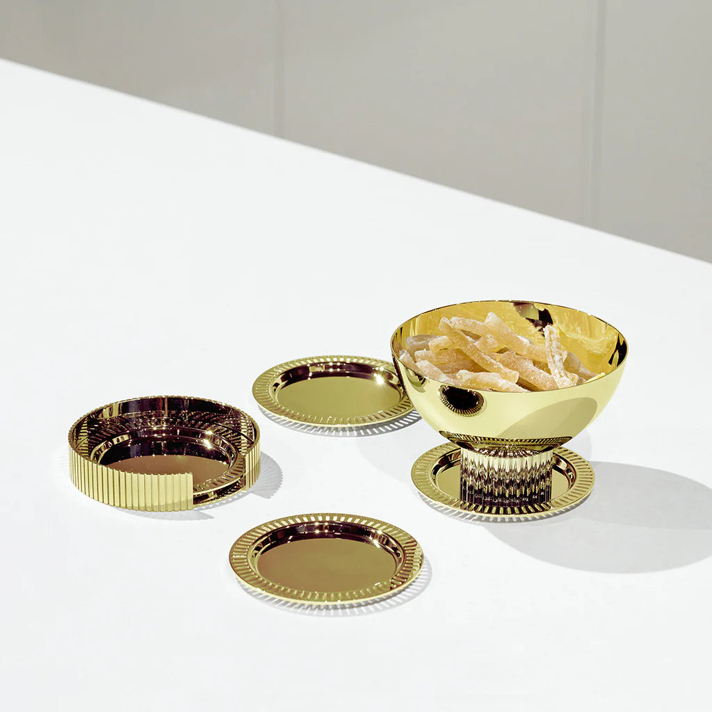 A shiny gold bowl filled with snacks rests on one of Anna New York’s Striations Coasters s/4 in Holder - Gold, accompanied by two additional empty Striations Coasters and a matching gold lid, all elegantly displayed on a white surface against a light background.