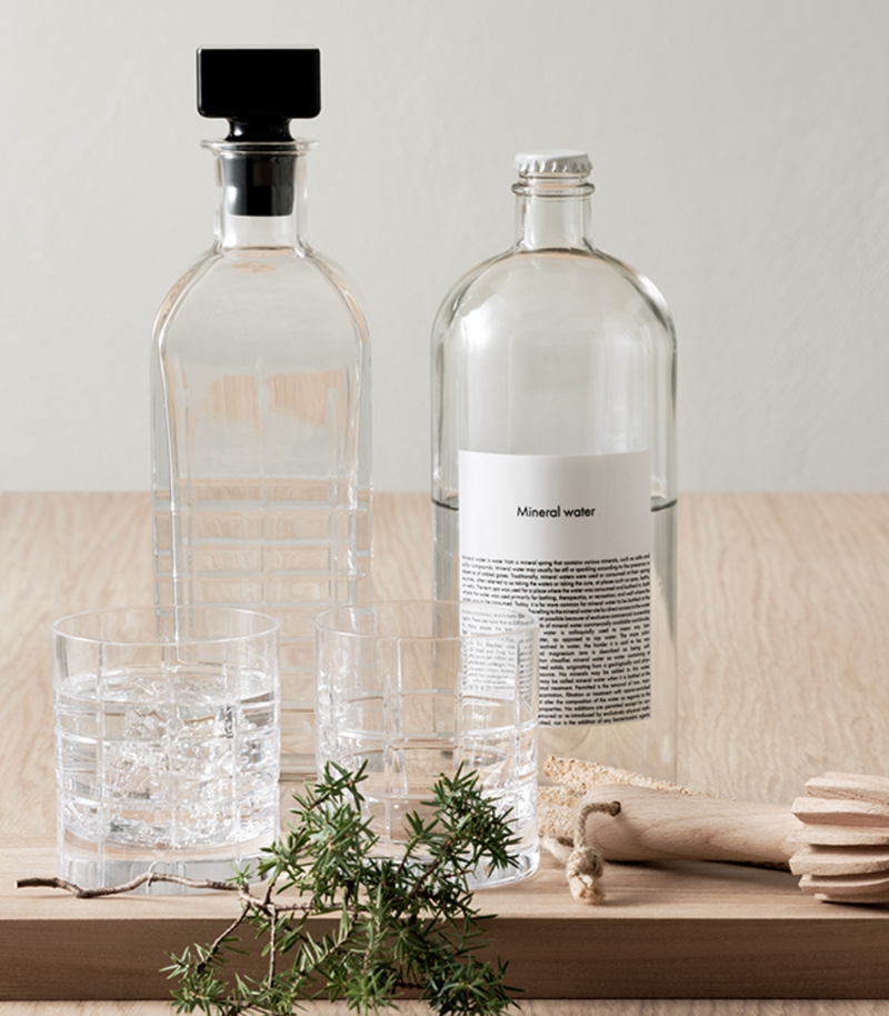 Two clear glass bottles (one labeled Mineral water) and an Orrefors Kosta Boda Street DOF Glass Set of 2, filled with water and ice, sit on a wooden surface alongside green herbs and wooden kitchen utensils.