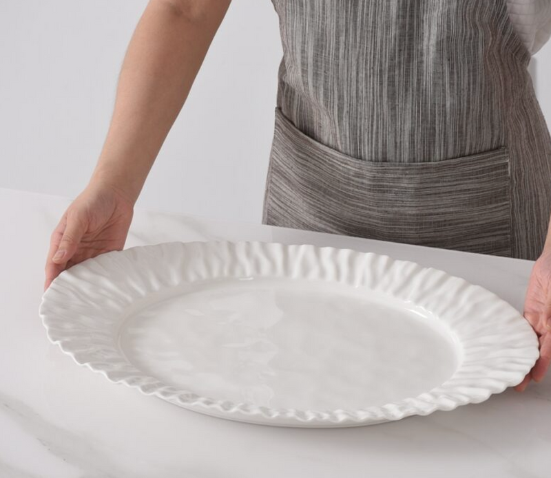 Wearing a striped gray apron, someone places the PAMPA BAY Mascali Bianca Serving Platter—microwave safe porcelain with wavy edges—on a white marble countertop.