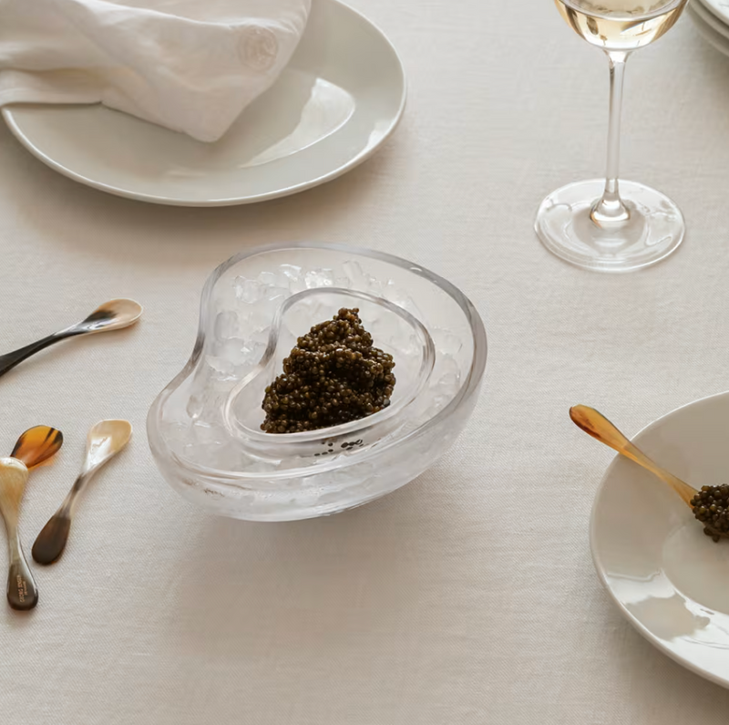 A table set with the Georg Jensen Indulgence Caviar Dish, a glass of white wine, assorted small spoons, a plate topped with a white napkin, and a glass bowl of ice and caviar on a crisp white tablecloth.