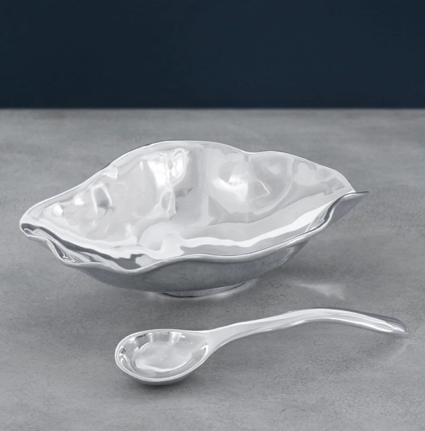The Beatriz Ball Vento Claire Bowl with Spoon, featuring a wavy, organic aluminum design and a matching curved-handled spoon, sits on a gray surface against a dark blue background.