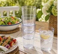 Four TarHong Provincial Jumbo Glasses (Clear 23oz) are stacked on an outdoor table, next to sparkling water with lemon, veggie skewers, salad, and white flowers. These glasses are shatterproof and dishwasher safe for easy cleaning.