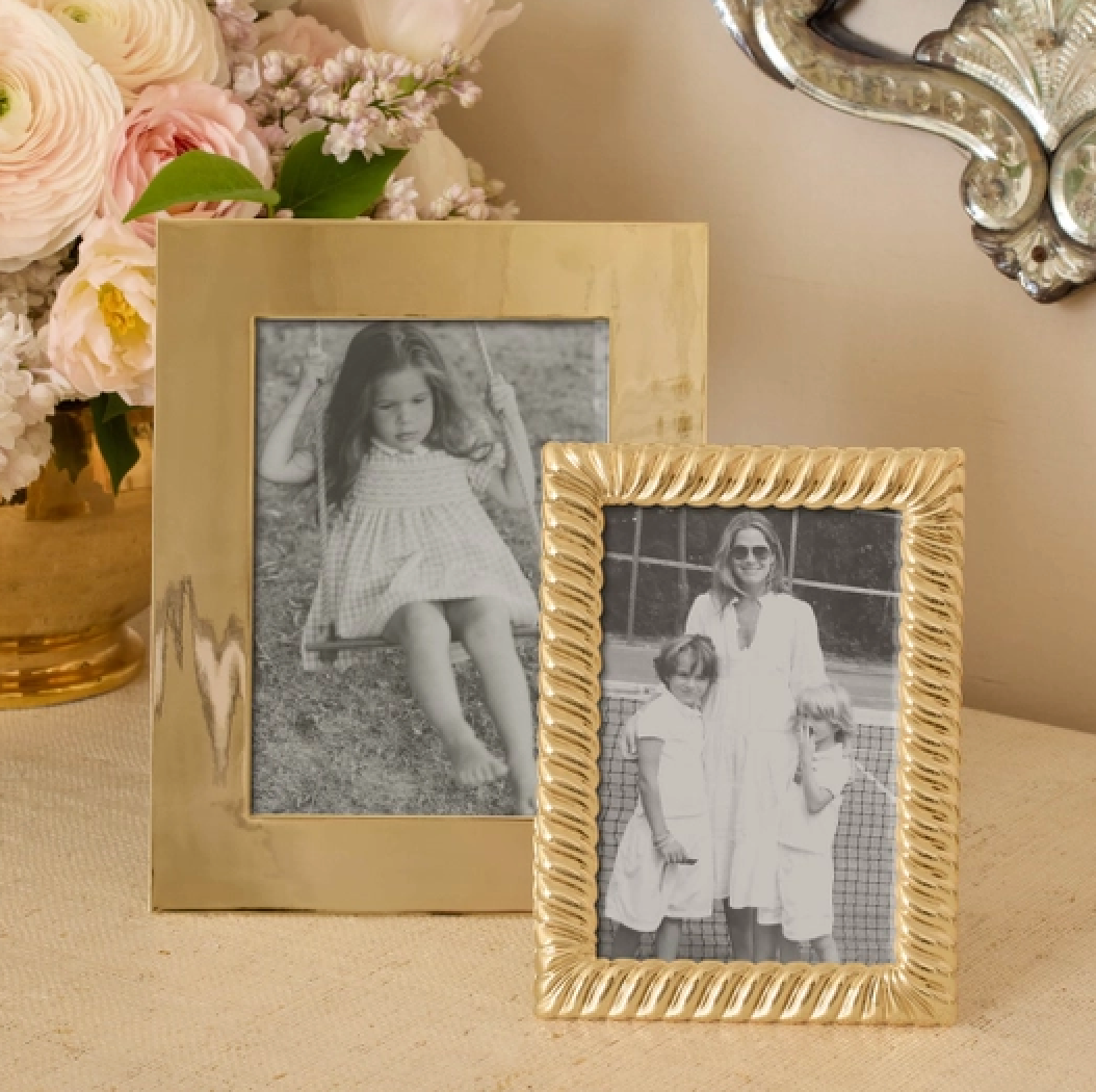 Two Loretta Frame Gold picture frames by Aerin sit on a table—one displays a 4x6 photo of a girl on a swing, the other shows a woman with two children. A vase of pink flowers and a decorative mirror are in the background.