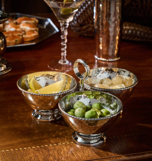 The Juliska Graham Triple Cocktail Bowl features three ornate hammered stainless steel bowls for garnishes—lemon wedges, green olives, and pearl onions—on a wooden table, with a martini glass and appetizer tray in the background.