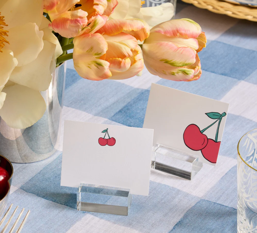 Two blank place cards with cherry illustrations stand in Dear Annabelle's Clara Place Card Holder Set of 6 - Clear on a blue and white checkered tablecloth, beside tulips in a silver vase, adding charm to this modern tablescape.
