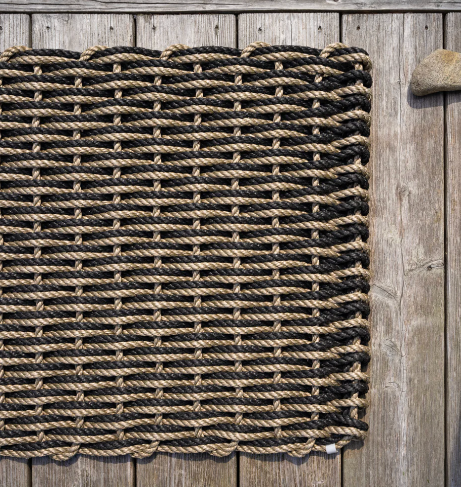 The Rope Co's Coastal Woven Outdoor Floormat in Sand & Charcoal (24 x 38) sits on a wooden deck, highlighting its durability with a large stone placed on the top right corner.
