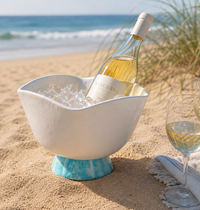 A bottle of white wine chills in the Beatriz Ball Resin Rio Wine Bucket White & Turquoise, set within a white bowl on a sandy beach with the ocean in the background. Two wine glasses and distinctive tableware, along with a white cloth, are arranged nearby to create an inviting seaside scene.