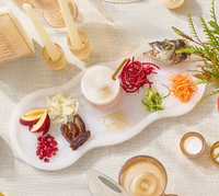 Celebrate Rosh Hashanah with the Waterdale Collection Resin Curved Simanim Tray Set, beautifully arranged with symbolic foods like apple slices, pomegranate seeds, dates, cabbage, pickled fish, shredded carrots, and greens. Displayed on a striking white tray and accented by candles and glassware atop a beige tablecloth, the festive ambiance is enhanced with a matching resin honey dish to sweeten your New Year celebrations.