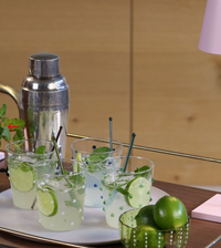 Six clear 15.2oz Party Dots Large Tumblers by Zafferano, each decorated with vibrant polka dots and filled with lime and mint cocktails topped with straws, sit on a white tray next to a cocktail shaker and a bowl of whole limes on a wooden table, bringing a cheerful touch to the scene.