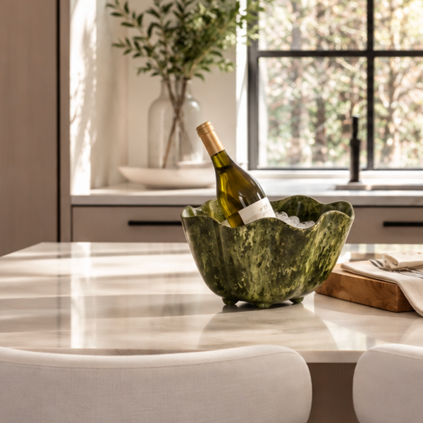 A bottle of white wine chills in the Beatriz Ball Resin Malachite Ice Bucket - Green, filled with ice and placed on a marble kitchen island. In the background, a vase of green branches sits near a sunlit window, while two white chairs are visible in the foreground.